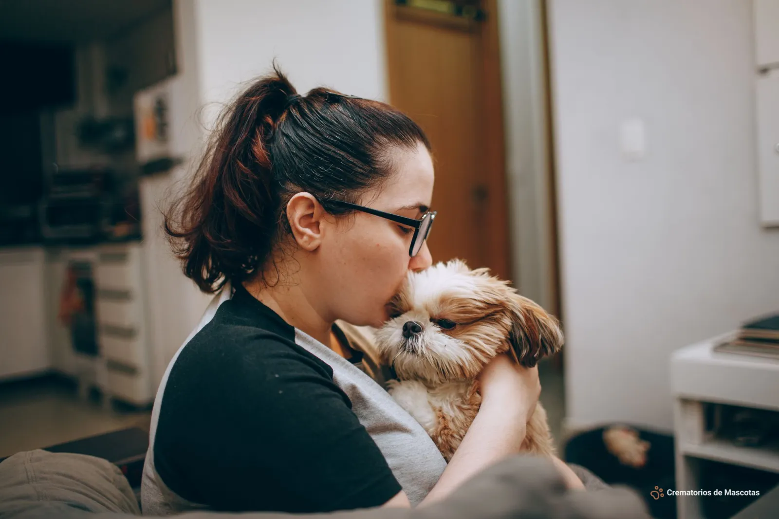 Una mujer con gafas besa con ternura a un perrito Shih Tzu de pelaje claro que sostiene en sus brazos